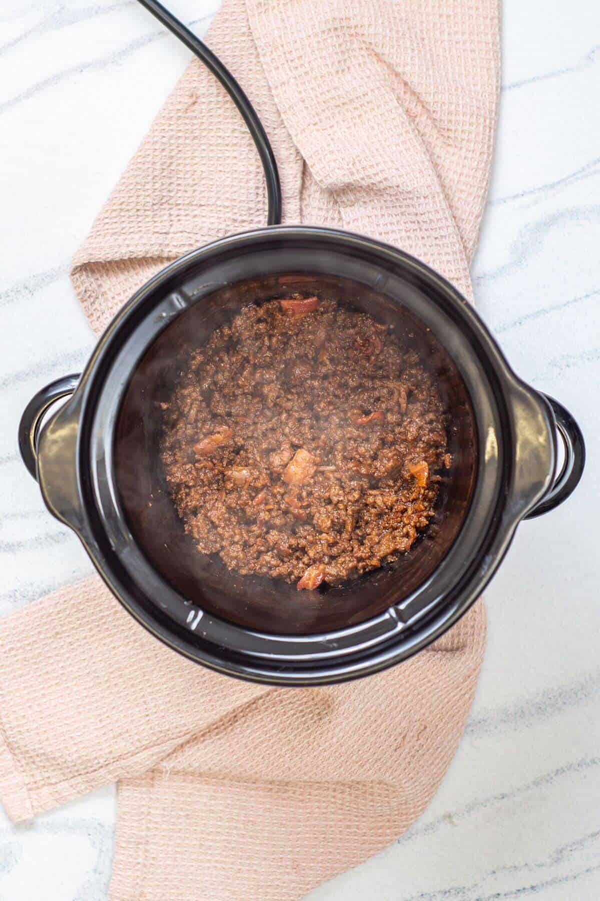 Overhead view of cooked ground meat in a black slow cooker, placed on a beige kitchen towel on a light countertop.