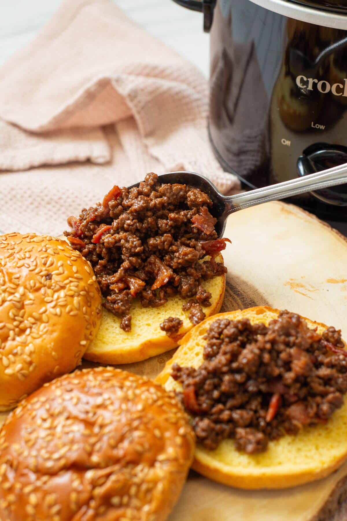 Spoonful of cooked ground meat mixture being placed onto an open sesame seed hamburger bun, with a slow cooker and another bun nearby.