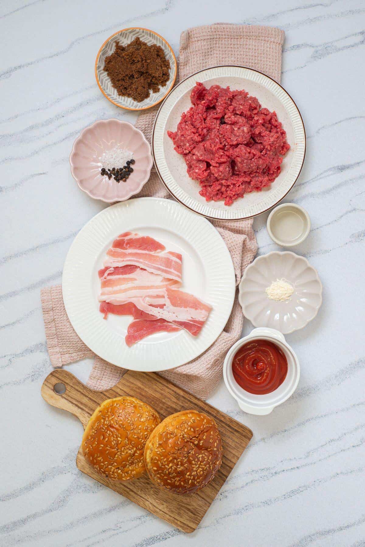 Burger ingredients arranged on a table, including ground beef, bacon, burger buns, ketchup, brown sugar, seasonings, and peppercorns in small bowls.