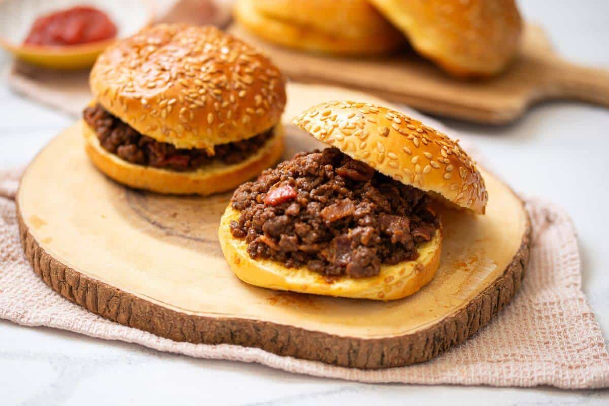 Two sesame seed buns filled with seasoned ground beef and diced tomatoes sit on a wooden board, with more buns in the background.