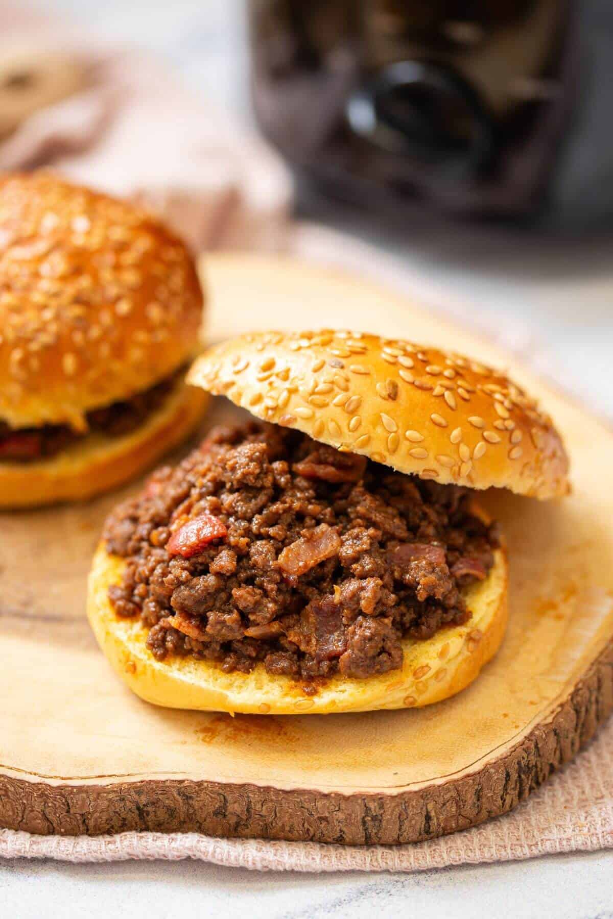 A sesame seed bun filled with seasoned ground beef sits open-faced on a wooden board, with another sandwich partially visible in the background.