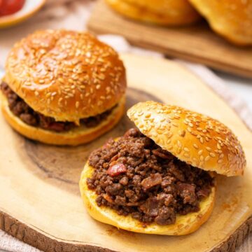 Two sesame seed buns filled with seasoned ground beef mixture, one sandwich open-faced, sitting on a wooden board.