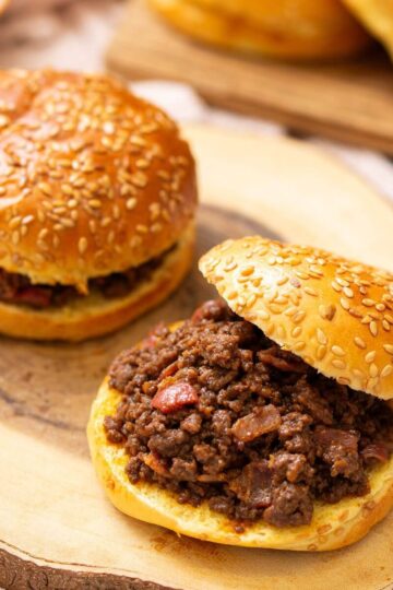 Two sesame seed buns filled with seasoned ground beef mixture, one sandwich open-faced, sitting on a wooden board.