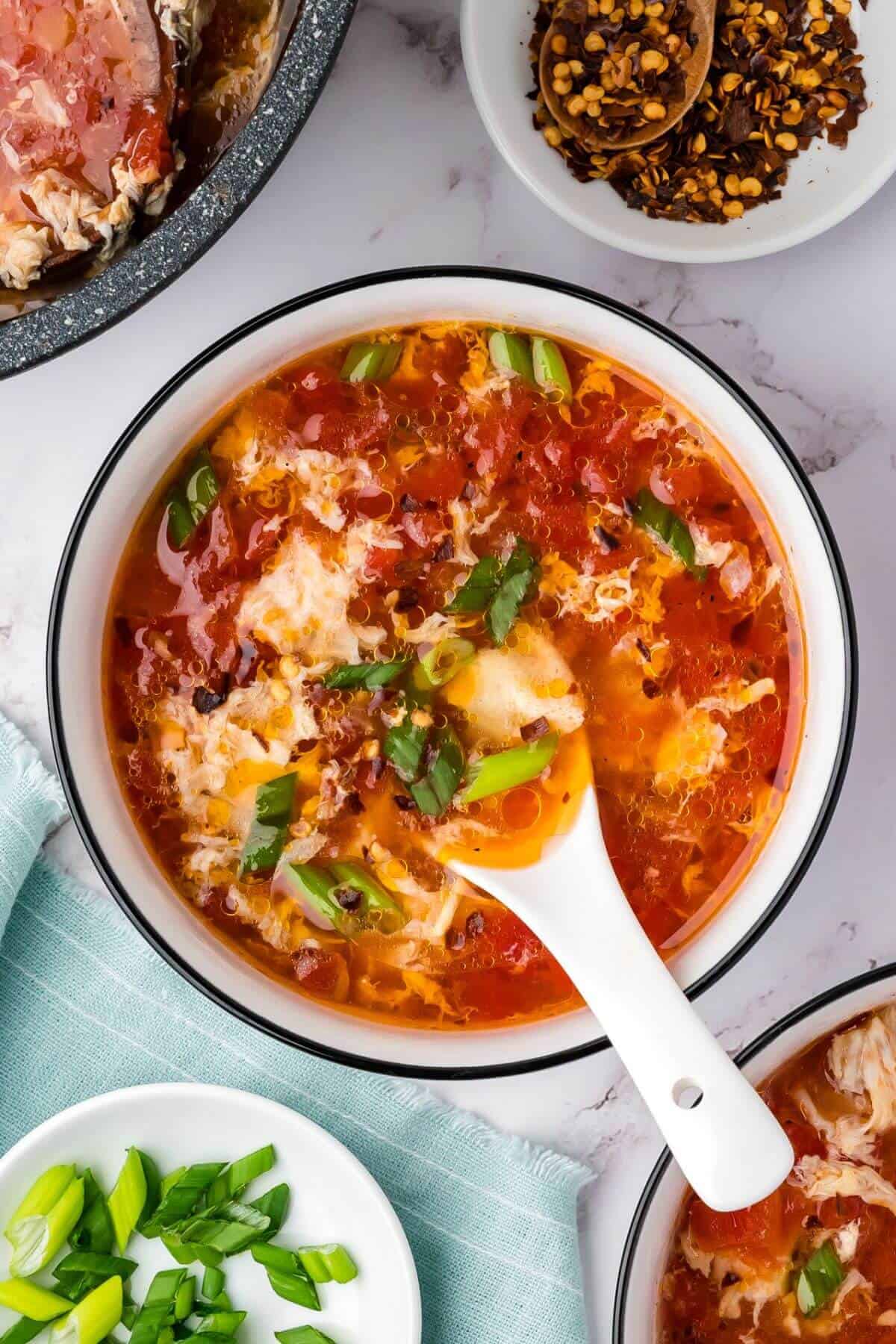A bowl of tomato egg drop soup garnished with chopped green onions, with a white spoon inside. Bowls of chili flakes and green onions are nearby on a marble surface.