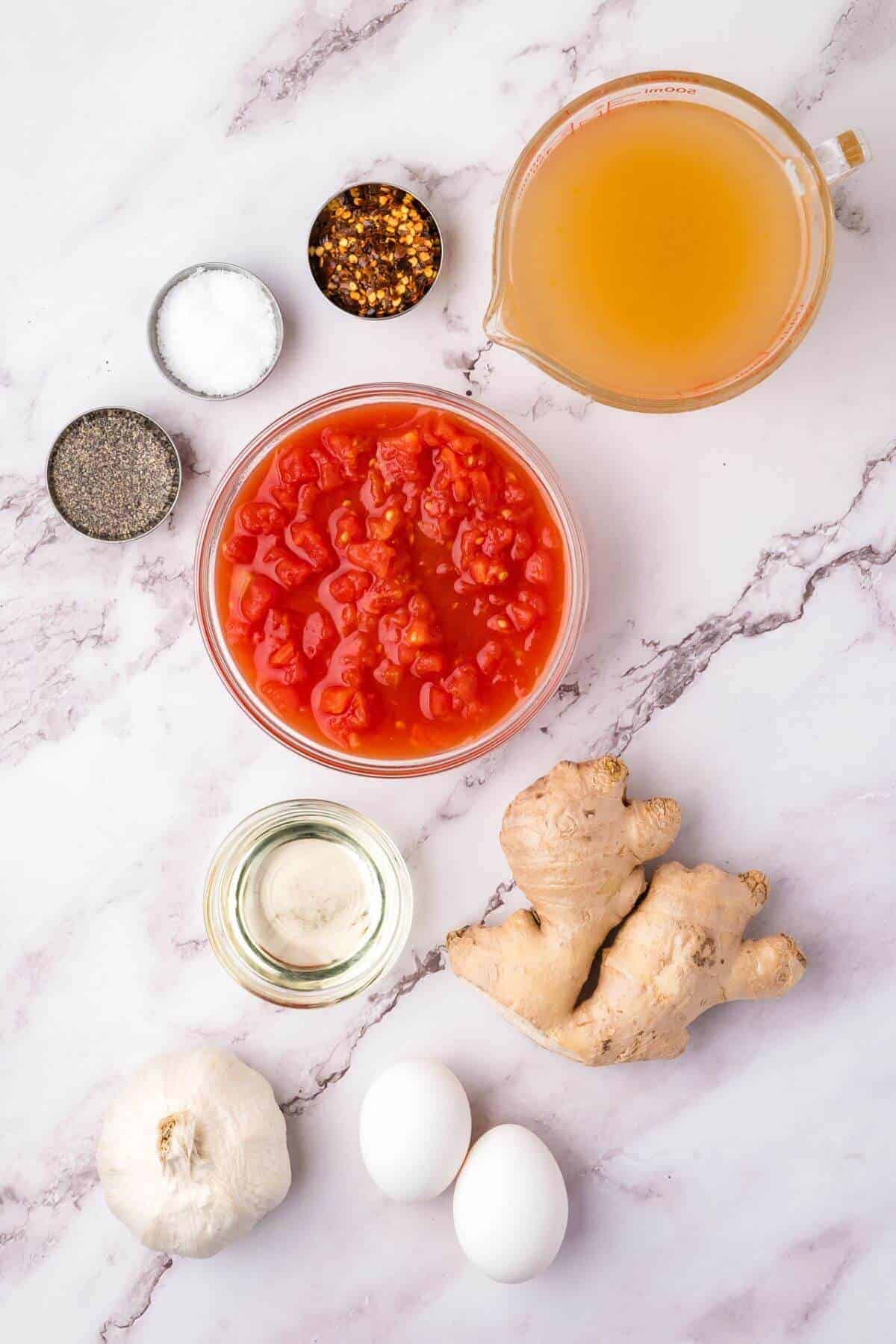 Assorted ingredients on a marble surface: diced tomatoes, ginger root, garlic bulb, two eggs, a measuring cup of broth, oil, salt, black pepper, and red pepper flakes in small bowls.