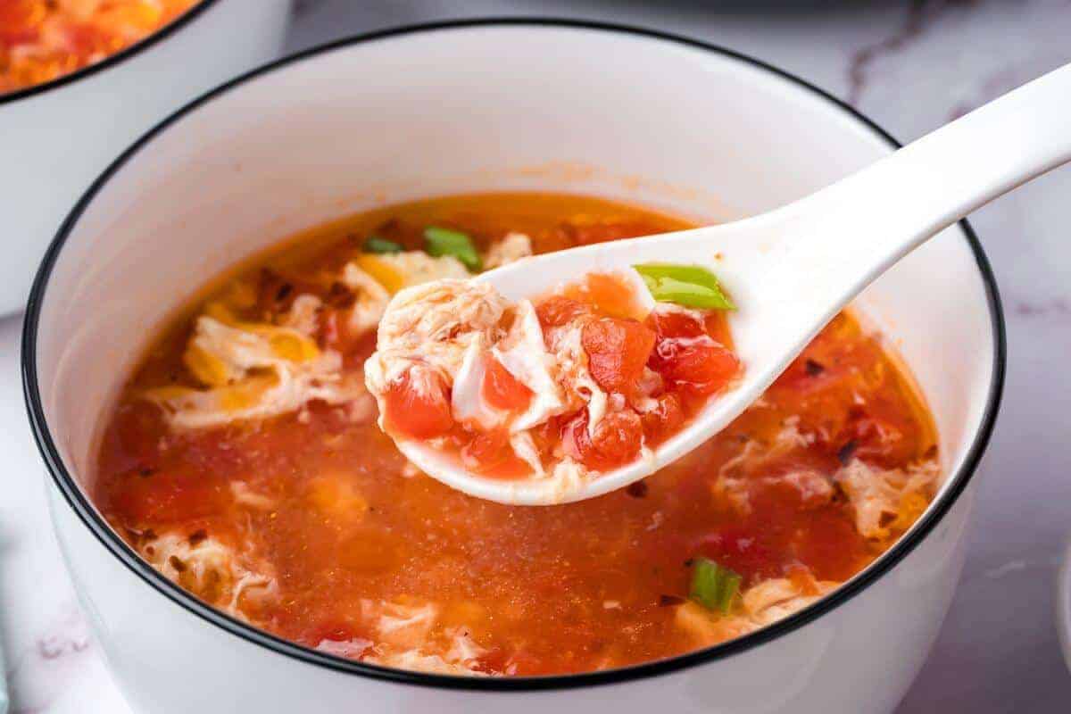 A close-up of a bowl of tomato egg drop soup, with a white spoon lifting a portion containing tomato chunks, egg ribbons, and green onions.