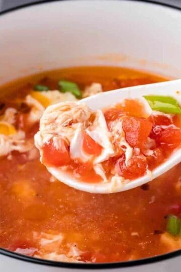 A white bowl of tomato egg drop soup with a ceramic spoon lifting a portion, showing diced tomatoes, egg ribbons, and green onions.
