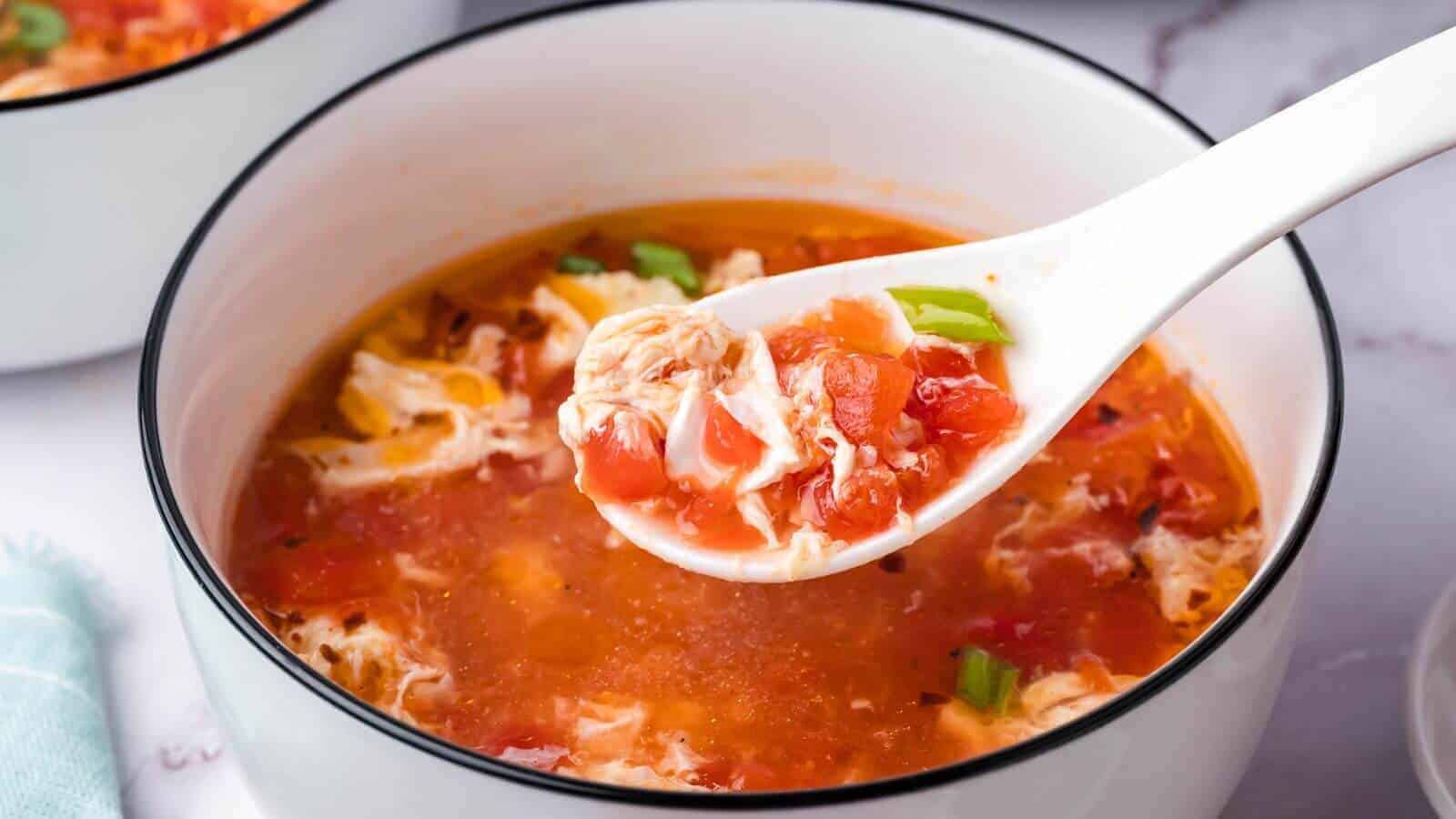 A white bowl of tomato egg drop soup with a ceramic spoon lifting a portion, showing diced tomatoes, egg ribbons, and green onions.