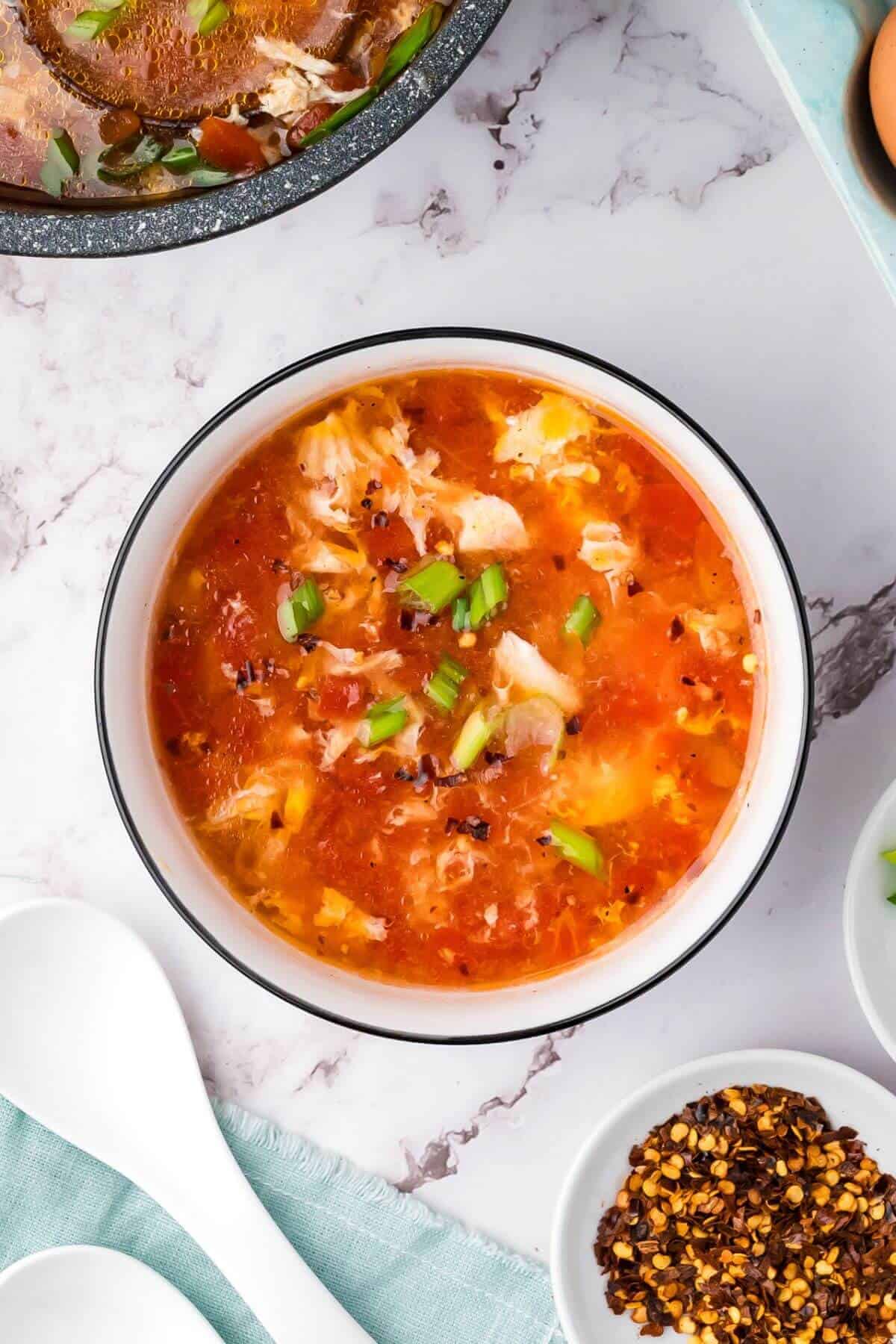 A bowl of hot and sour soup garnished with chopped green onions, with chili flakes and soup spoons nearby on a marble surface.