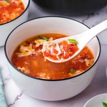 A white bowl filled with tomato egg drop soup, with a spoon lifting some soup, showing tomato and egg strands.