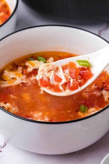 A white bowl filled with tomato egg drop soup, with a spoon lifting some soup, showing tomato and egg strands.
