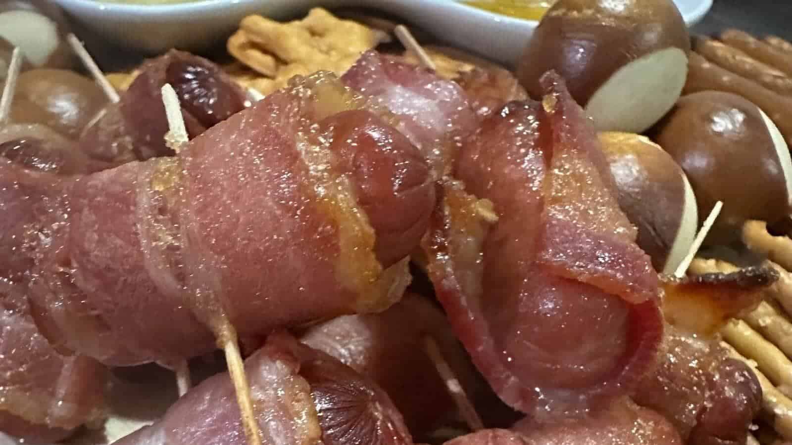 Close-up of bacon-wrapped sausages secured with toothpicks, surrounded by pretzel buns, crackers, and dipping sauces in the background.