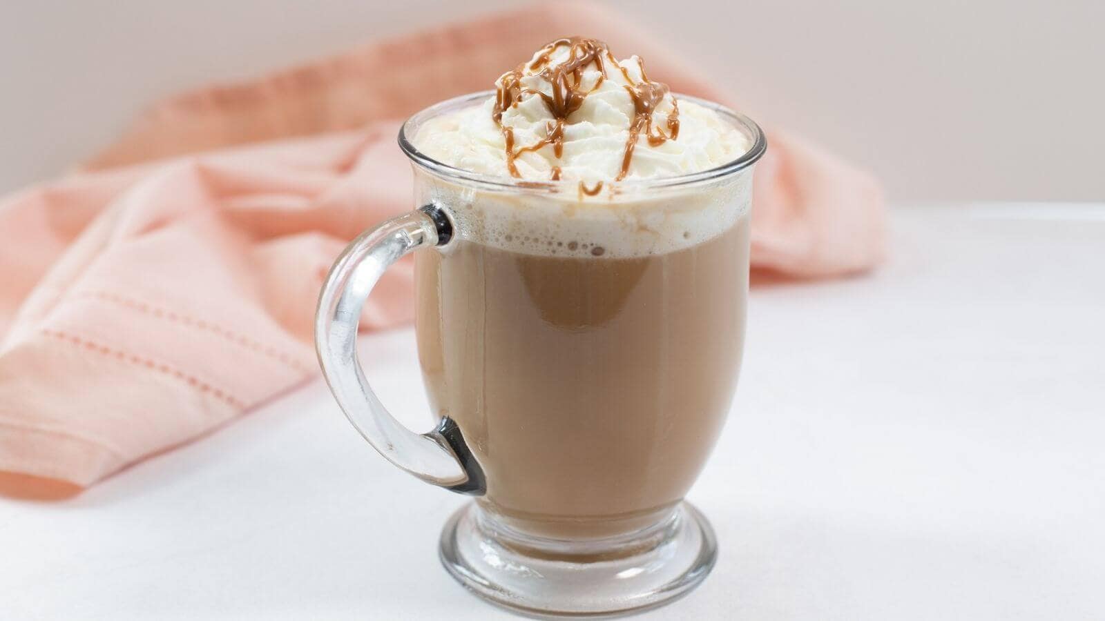 A glass mug of coffee topped with whipped cream and caramel drizzle sits on a white surface with a pink cloth in the background.
