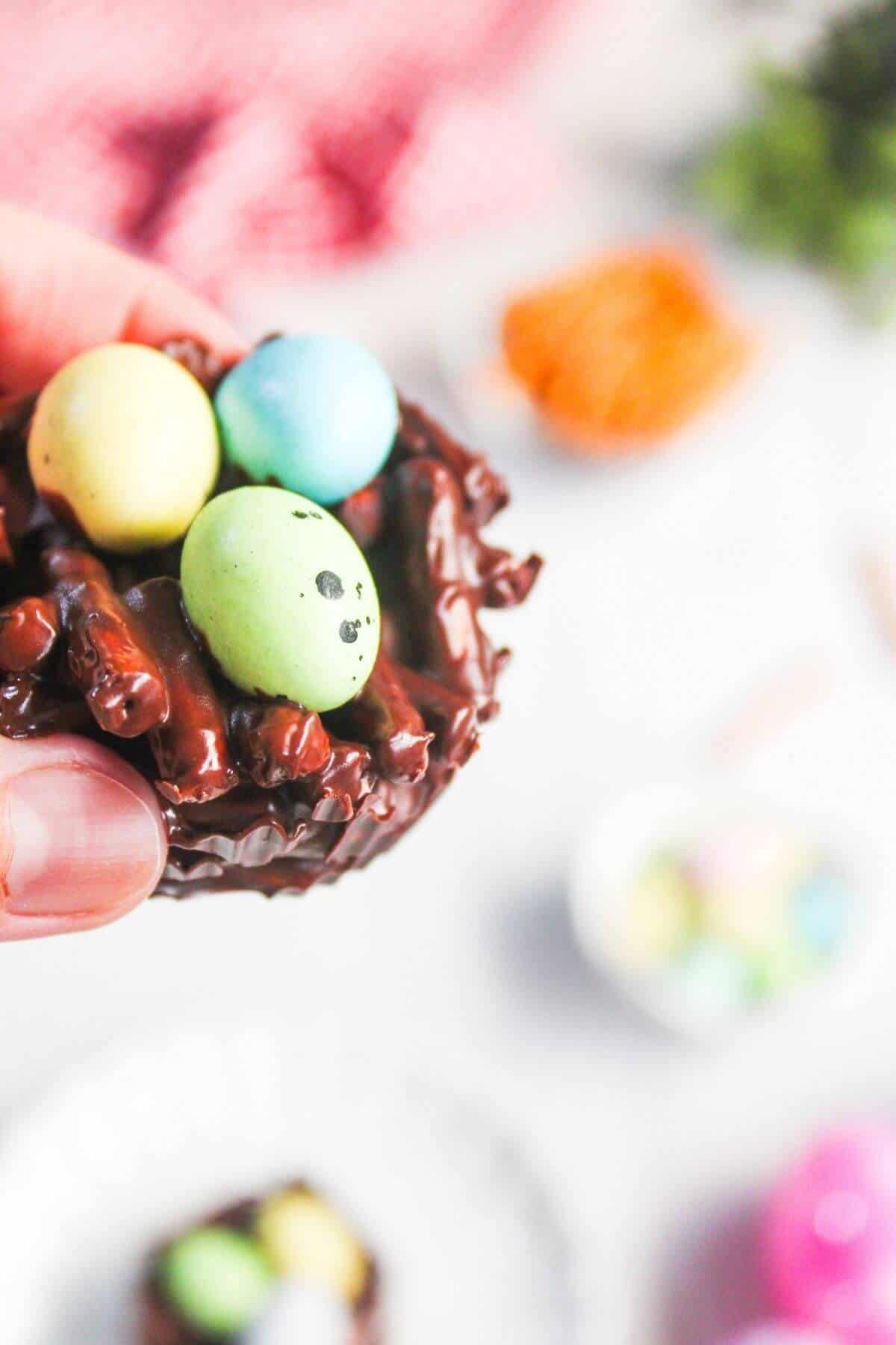 A hand holds a chocolate nest topped with three candy eggs, one yellow, one green with dots, and one blue, against a blurred background with treats.