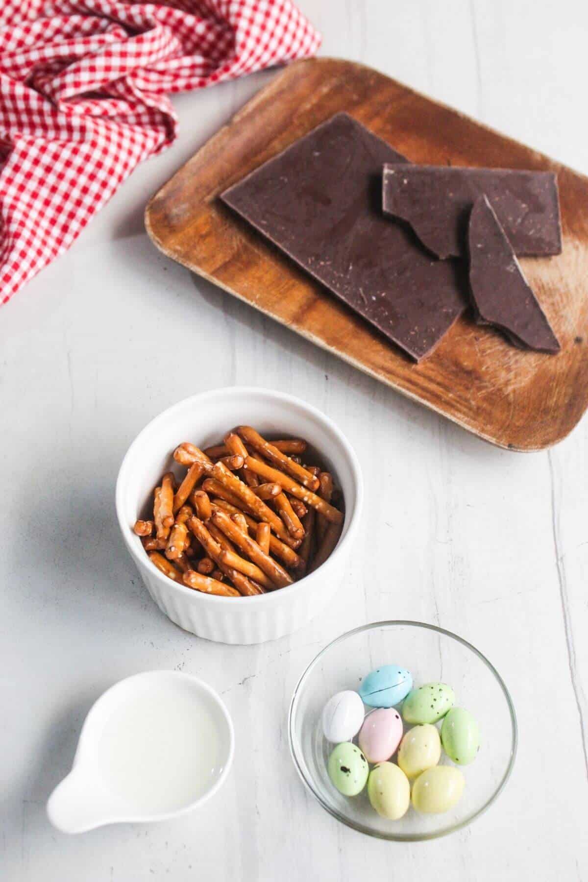 A wooden tray with chocolate pieces, a ramekin of pretzel sticks, a small pitcher of milk, and a bowl of pastel candy eggs on a white surface with a red checkered cloth.
