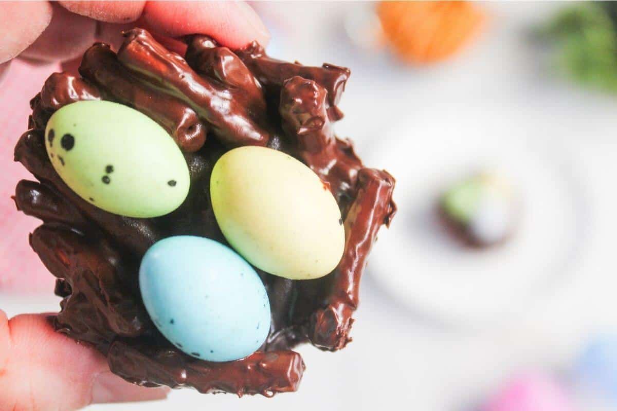 A hand holds a chocolate nest made of chocolate-covered pretzel sticks, with three pastel-colored candy eggs inside.