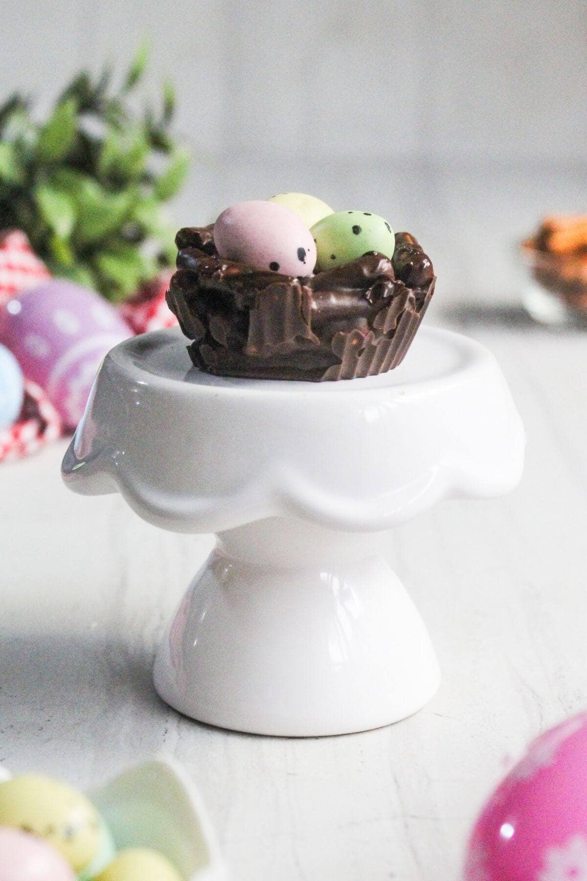 A chocolate nest with pastel candy eggs on top sits on a small white pedestal stand.