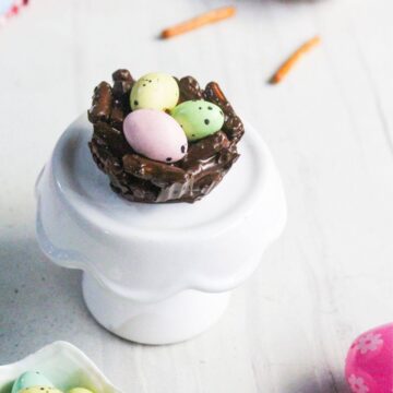 A chocolate nest made of pretzel sticks holds three pastel candy eggs, displayed on a white pedestal against a light background.