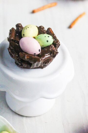 A chocolate nest made of pretzel sticks holds three pastel candy eggs, displayed on a white pedestal against a light background.