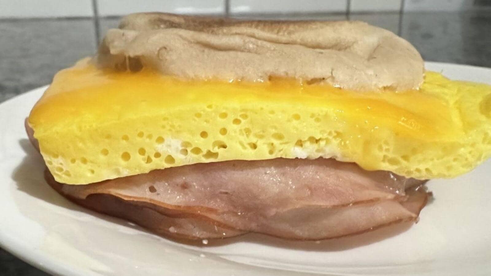 A breakfast sandwich with a slice of ham, a layer of cooked egg, and an English muffin top on a white plate.