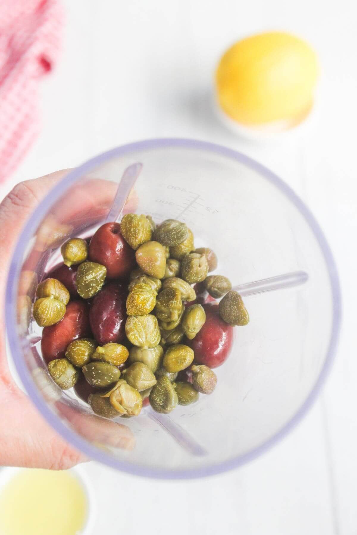 A hand holds a blender cup containing green capers and red olives, with a lemon and a bowl of liquid visible in the background.