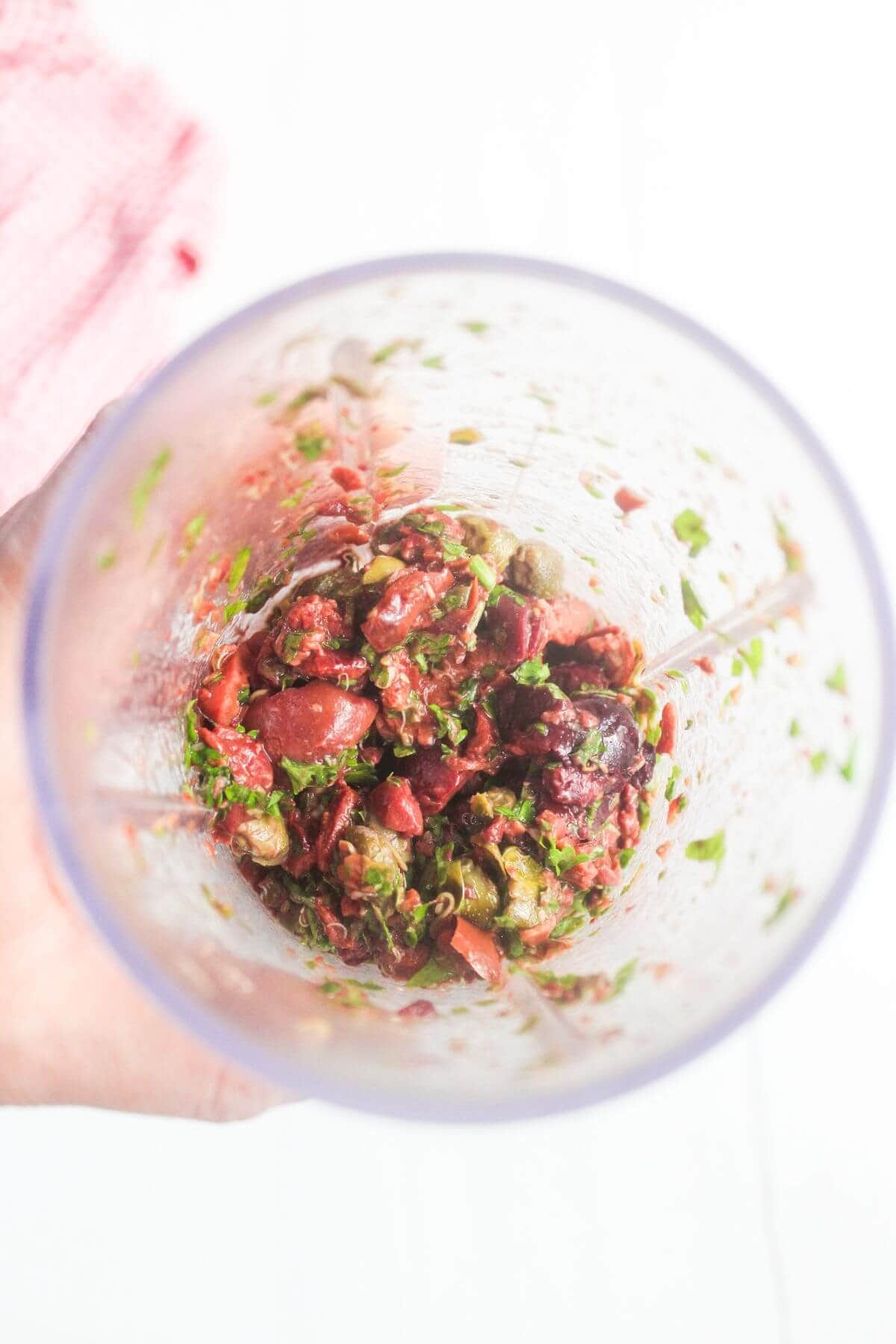 Top view of a blender cup containing a roughly blended mixture of olives, herbs, and capers, held by a hand against a white background.