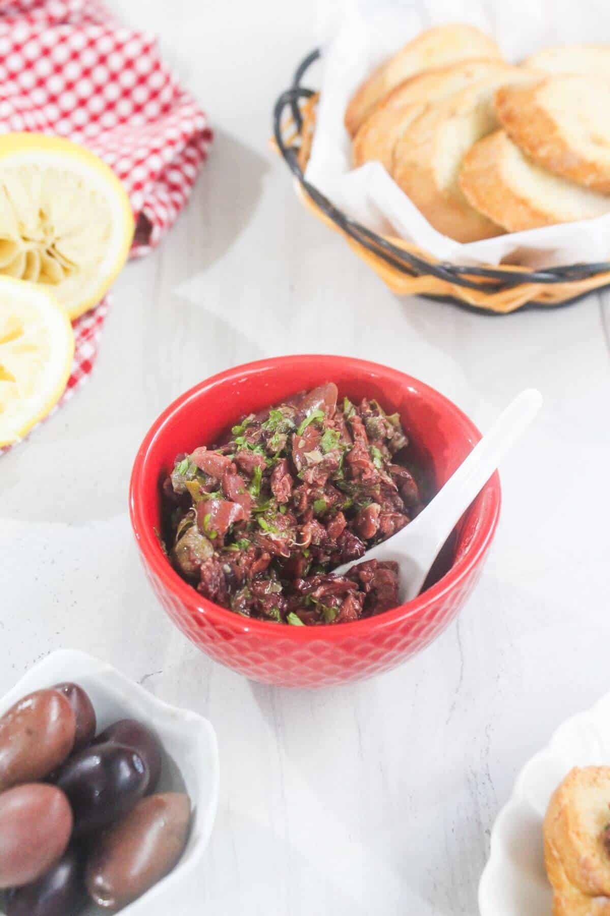 A red bowl of chopped olive tapenade with a spoon, surrounded by a basket of toasted bread slices, whole olives, and lemon halves on a white surface.