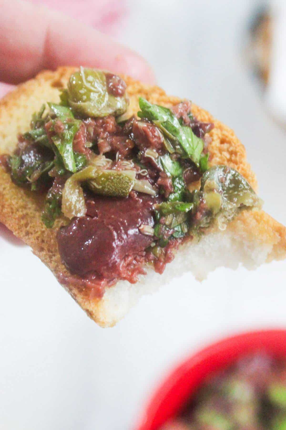 A hand holding a piece of toasted bread topped with olive tapenade, chopped herbs, and visible green and black olives.