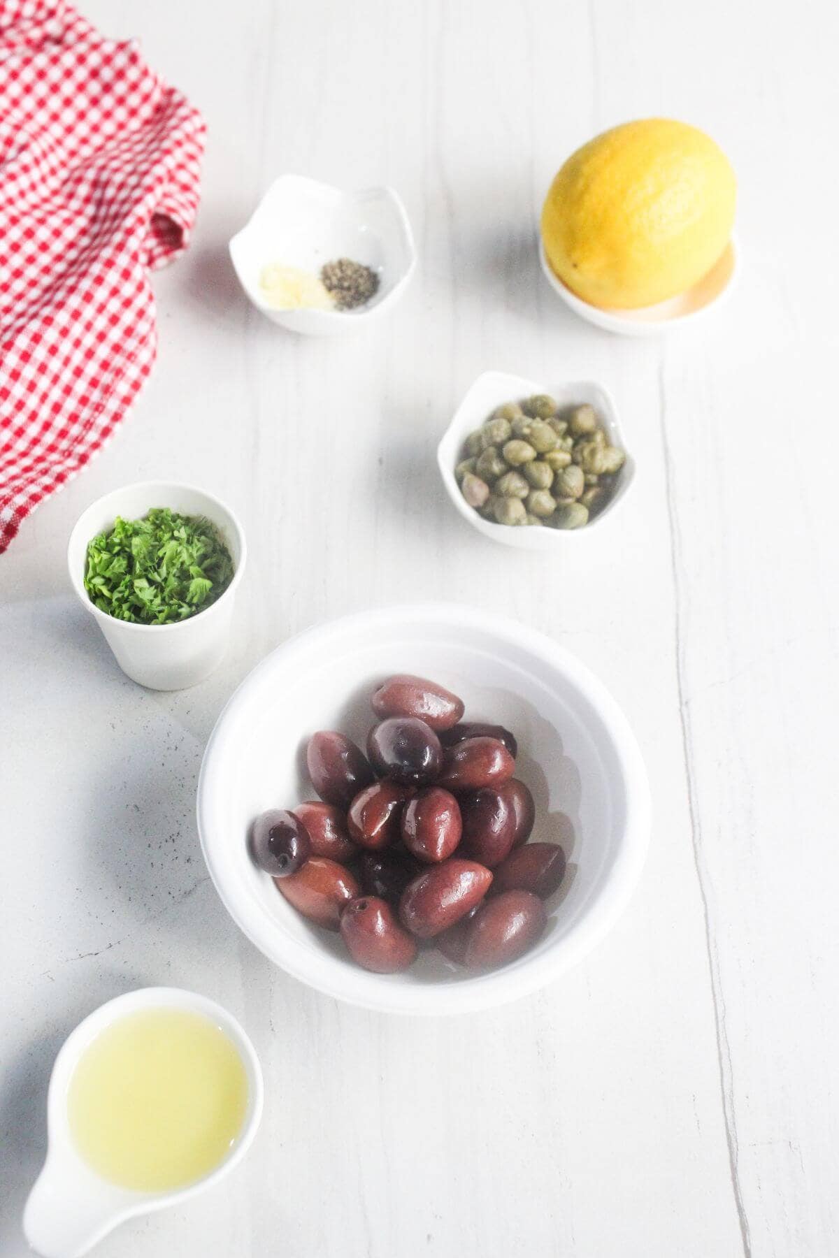 A bowl of Kalamata olives, a lemon, capers, chopped herbs, lemon juice, and spices on a white surface with a red checkered cloth.