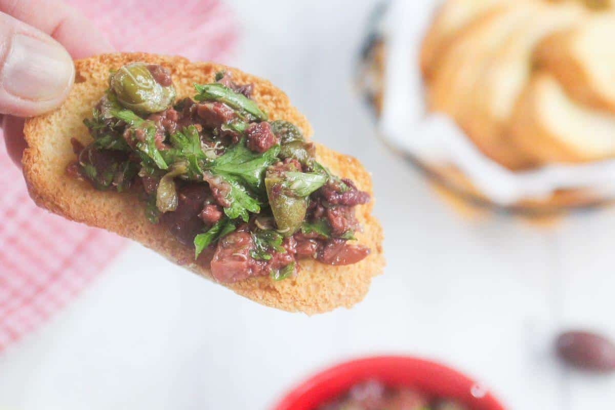 A hand holding a slice of toasted bread topped with olive tapenade, with a bowl of bread and a red dish in the background.