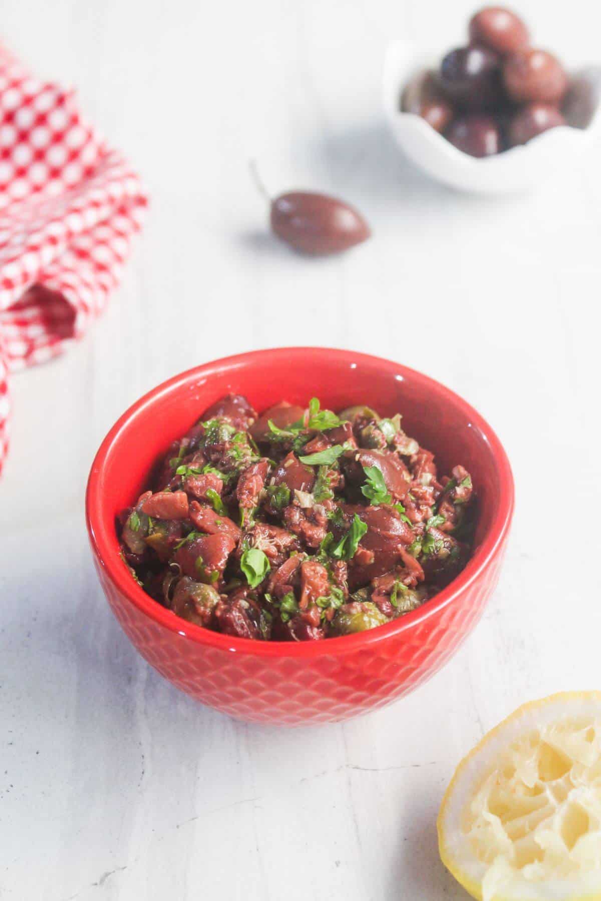 A red bowl filled with chopped olive tapenade garnished with herbs sits on a white surface, with a squeezed lemon half and whole olives in the background.