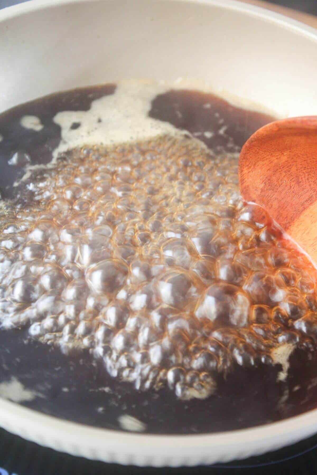 A close-up of a pan with dark liquid boiling and bubbling, being stirred with a wooden spoon.