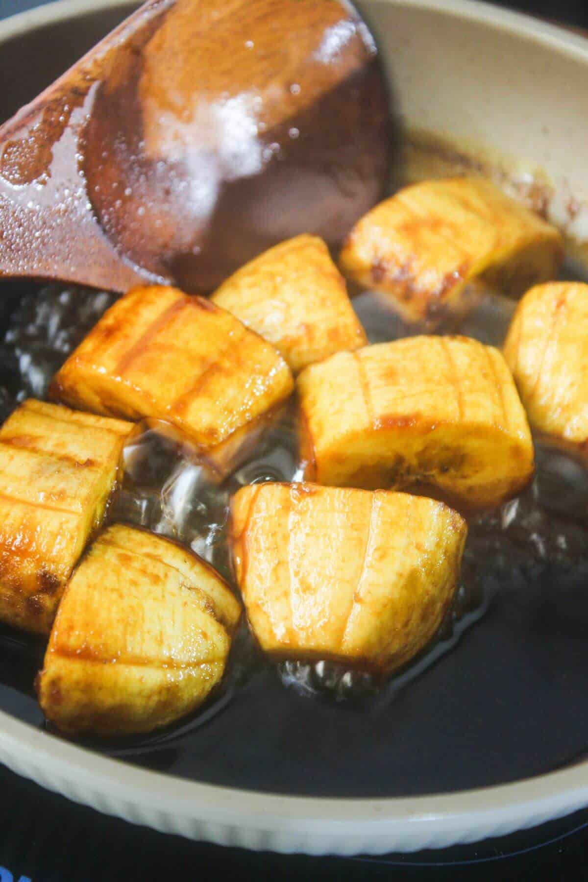 Sliced plantains frying in oil in a pan with a wooden spatula.