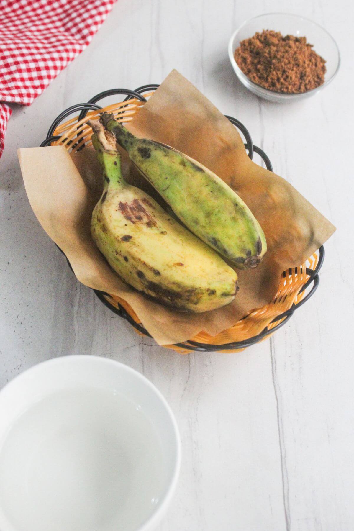 A basket with two green plantains on brown paper, a bowl of water, and a small bowl of brown sugar on a white surface.