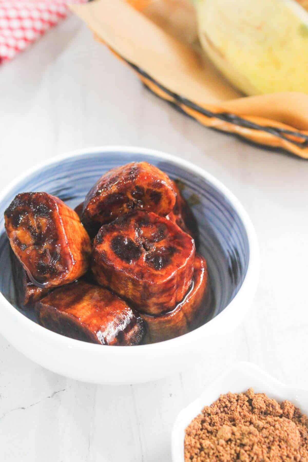 A white bowl filled with caramelized, sliced plantains on a light-colored surface with sugar and a banana visible in the background.