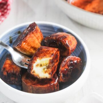 A bowl of caramelized plantain slices with a fork picking up one piece showing the soft inside.
