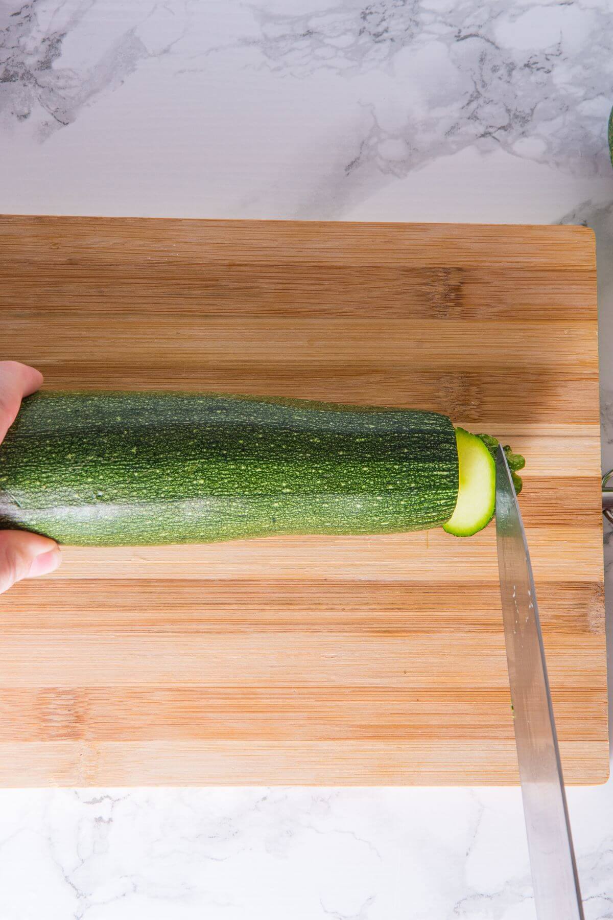 A person is slicing the end off a zucchini on a wooden cutting board with a knife.