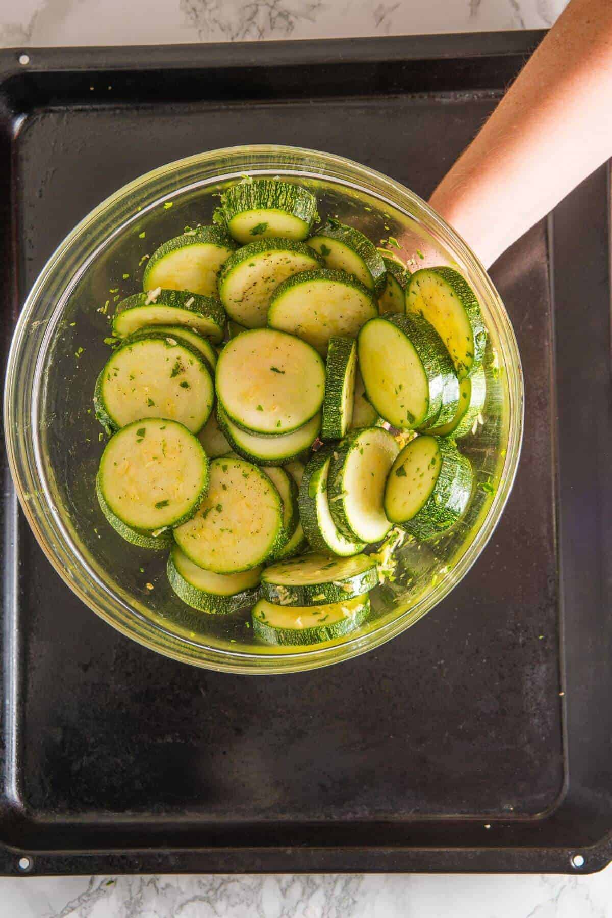 A glass bowl of sliced zucchini mixed with herbs and oil is held above a black baking tray.