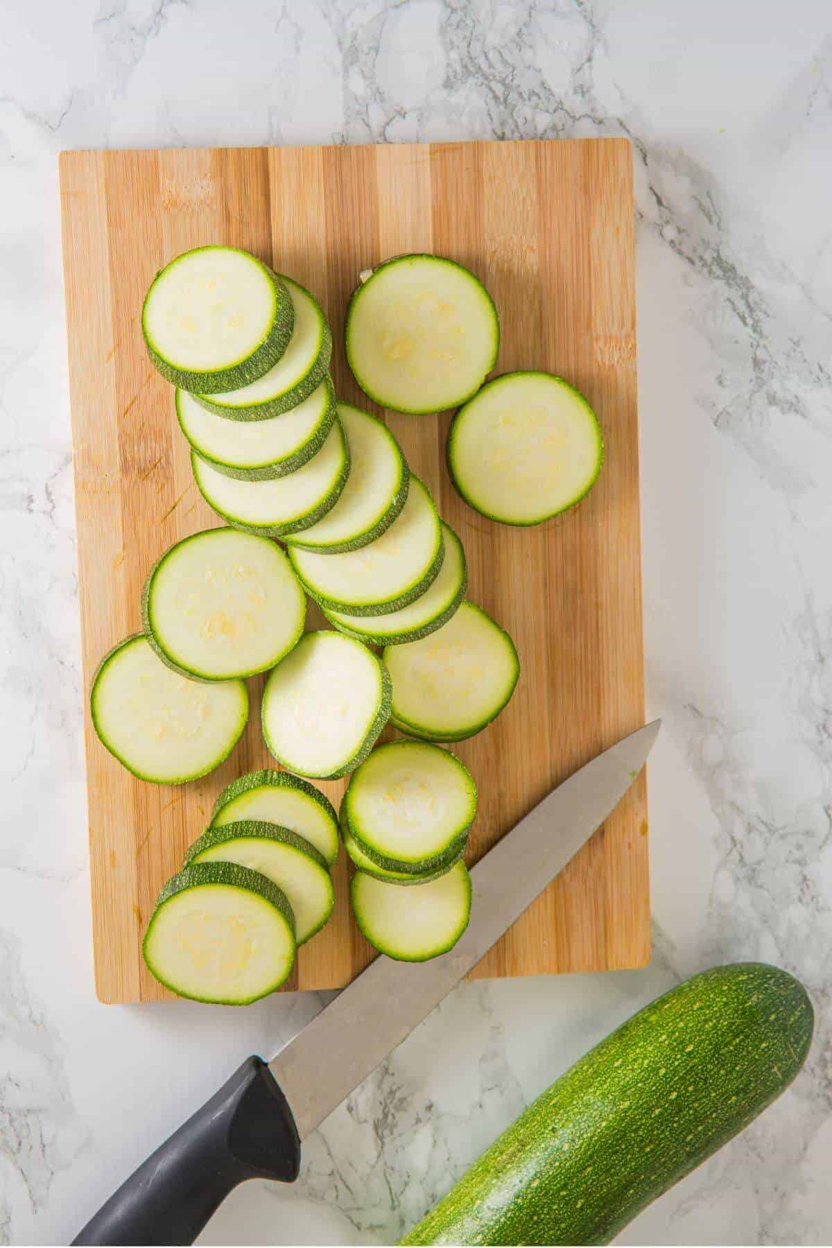 Sliced zucchini arranged on a wooden cutting board with a knife and a whole zucchini on a marble surface.