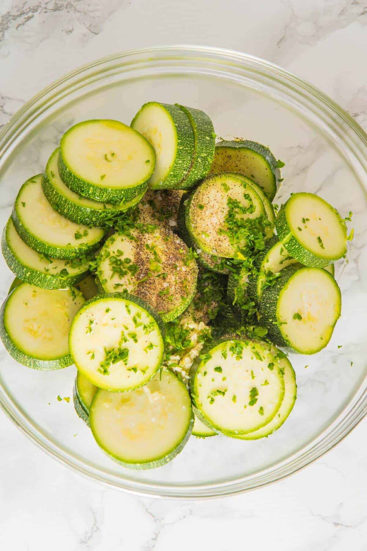 Sliced zucchini in a glass bowl, topped with chopped herbs, ground pepper, and seasonings on a white marble surface.