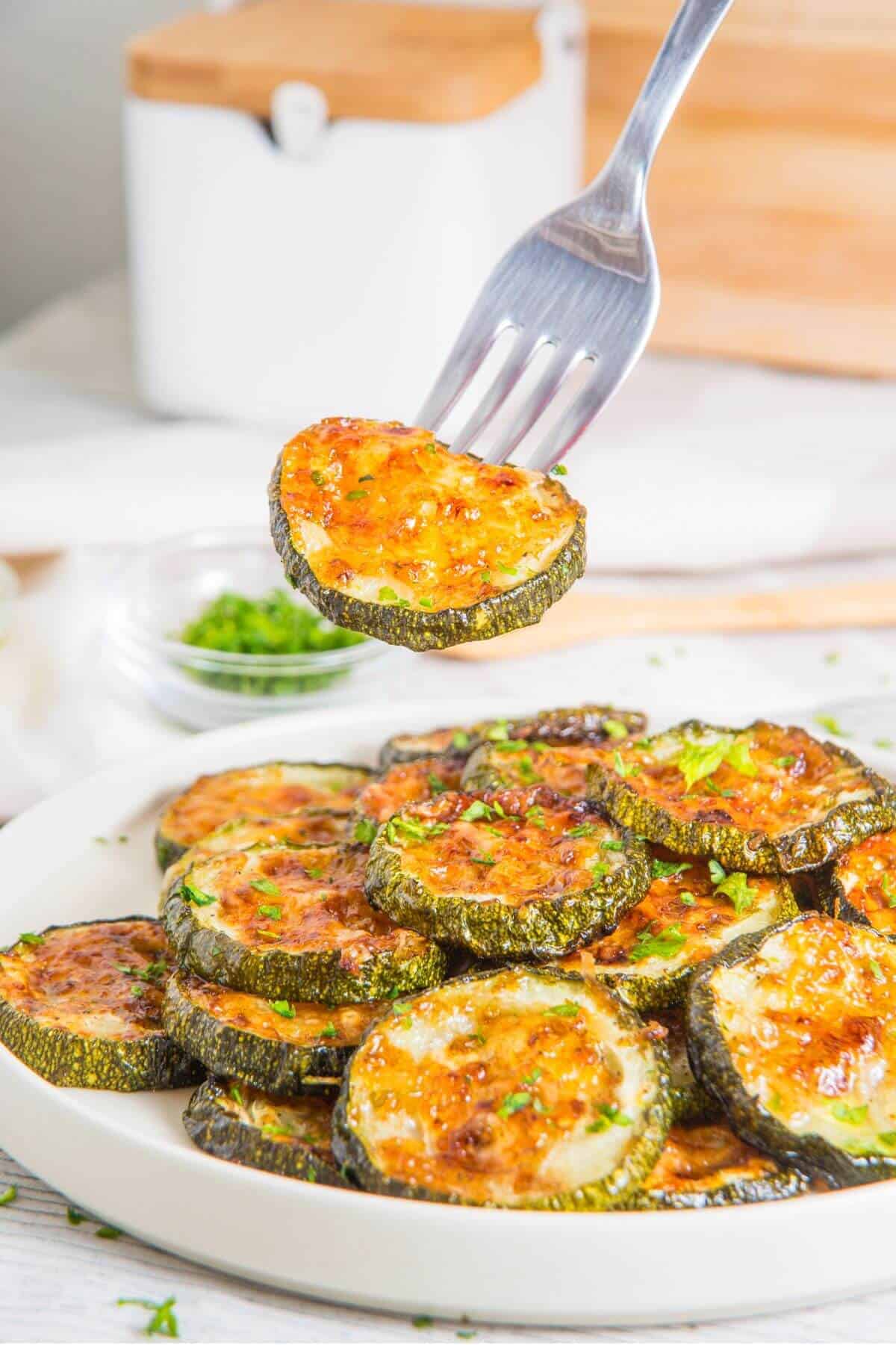 A plate of baked zucchini slices topped with melted cheese, with a fork holding one slice above the plate.