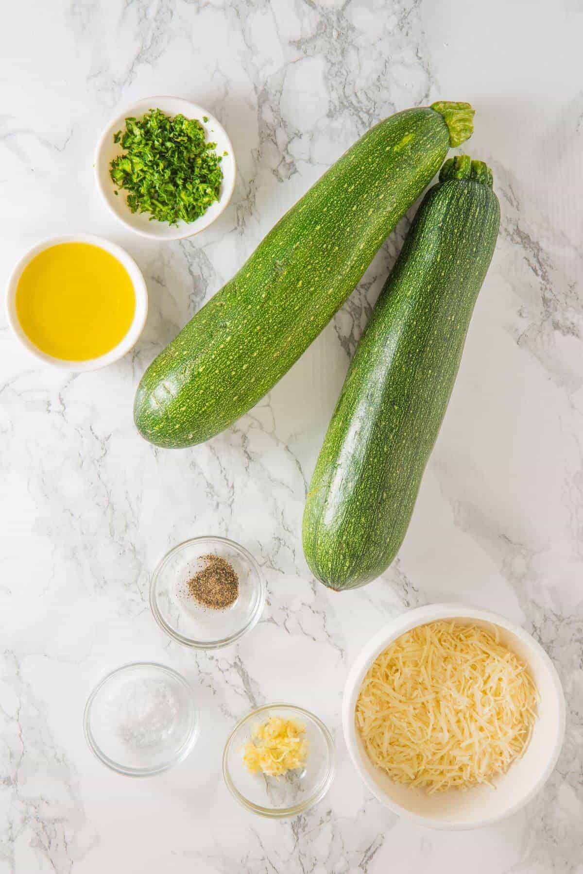 Two whole zucchinis, grated cheese, olive oil, chopped herbs, salt, pepper, and minced garlic arranged on a white marble surface.