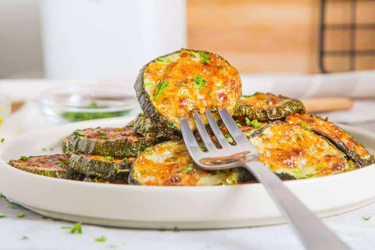 A fork holds a slice of baked zucchini topped with melted cheese and herbs, above a plate stacked with more zucchini slices.
