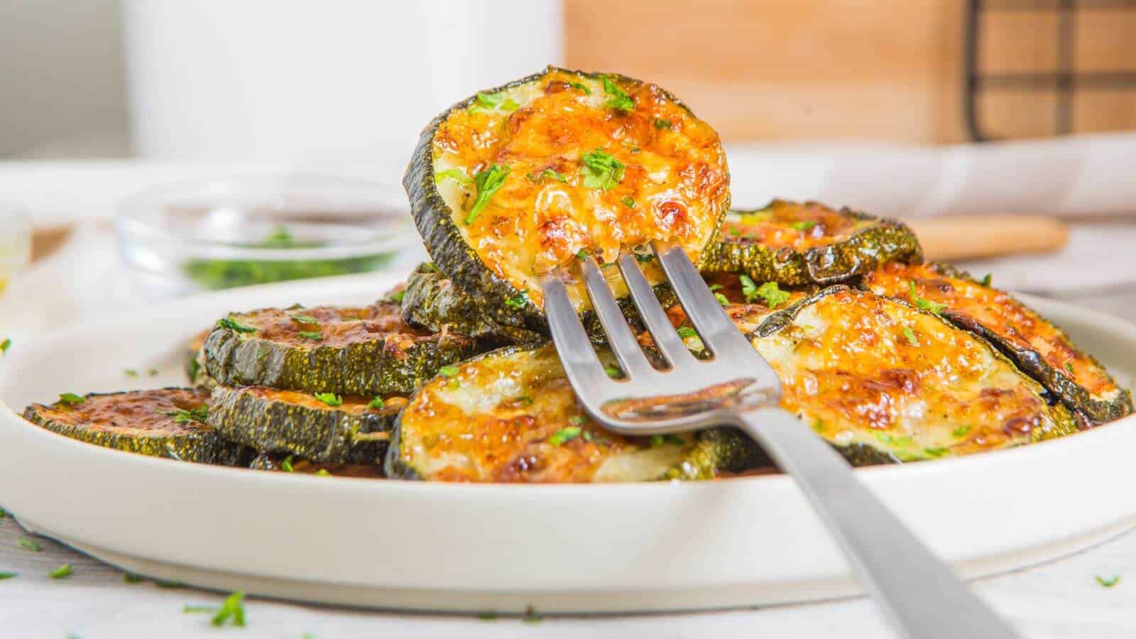A fork holding a slice of baked zucchini with melted cheese on top, resting on a plate with more cheesy zucchini slices.