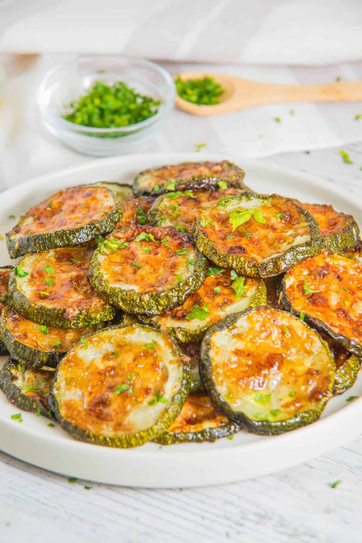 A plate of baked zucchini slices topped with melted cheese and herbs, with bowls of chopped herbs in the background.