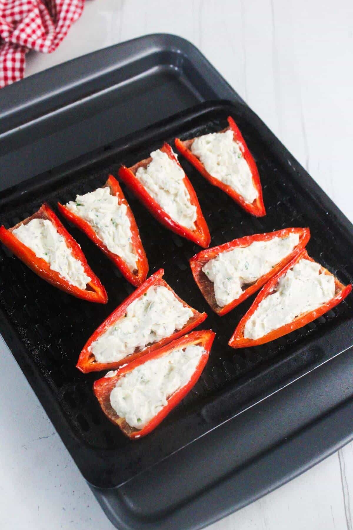 Halved red peppers filled with a creamy white cheese mixture are arranged on a black grilling tray, ready to be cooked.