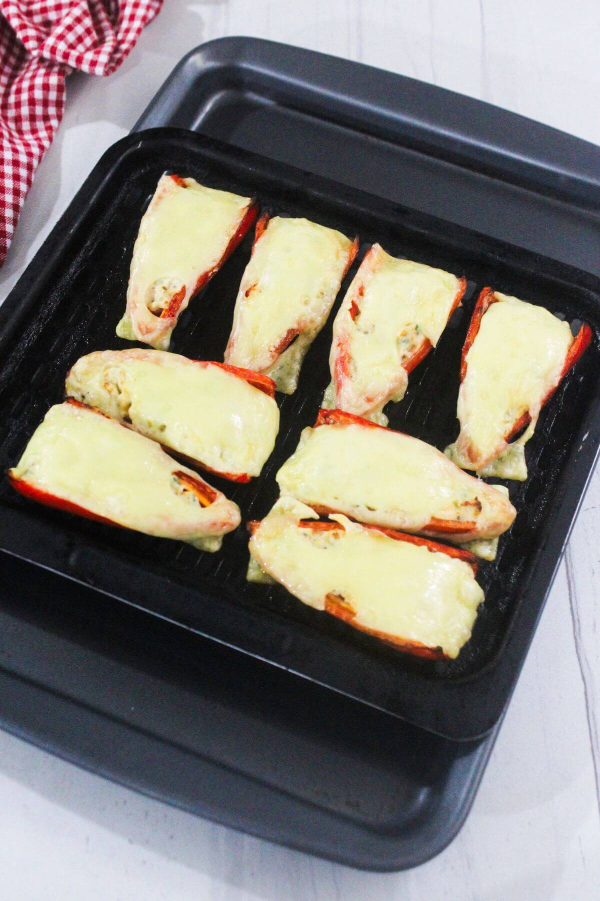 Eight stuffed red bell pepper halves topped with melted cheese are arranged on a black baking tray.