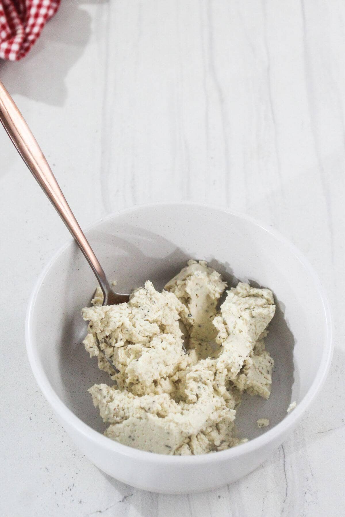 A white bowl containing a chunky white mixture with herbs, with a spoon resting inside, placed on a light marble surface.