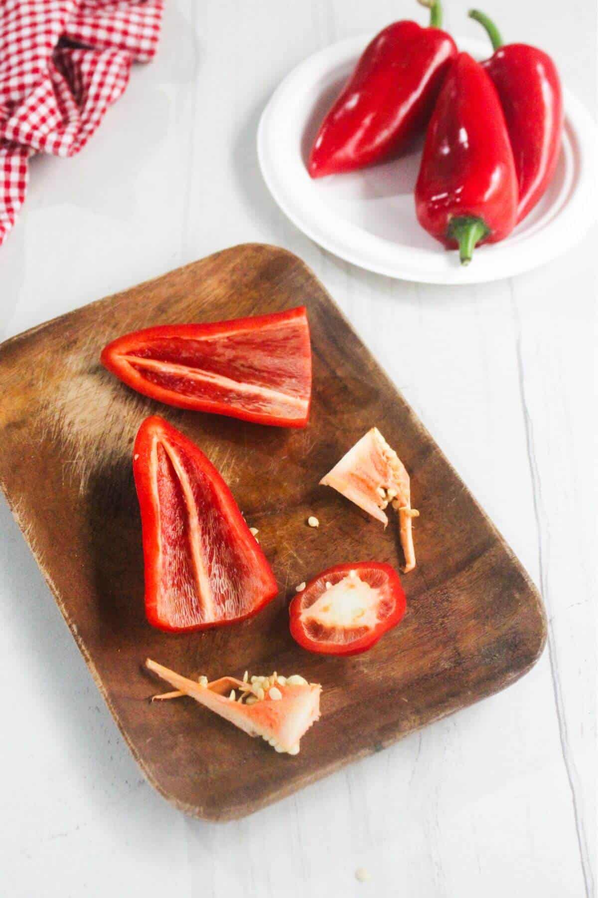 A wooden board with a sliced red pepper, seeds, and stems; a plate with whole red peppers is in the background.