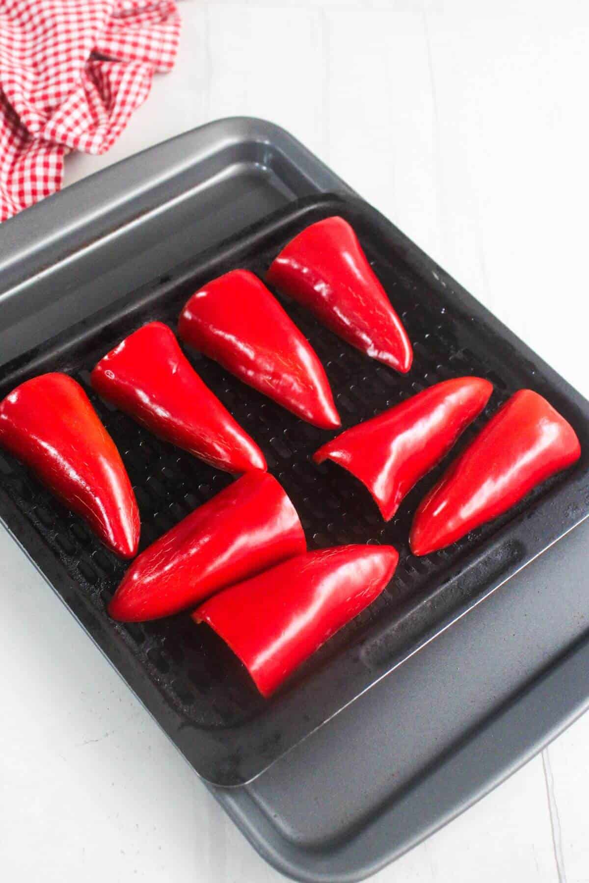 Eight halved red bell peppers are arranged on a black grill tray, ready to be roasted. A red and white checkered cloth is visible in the corner.